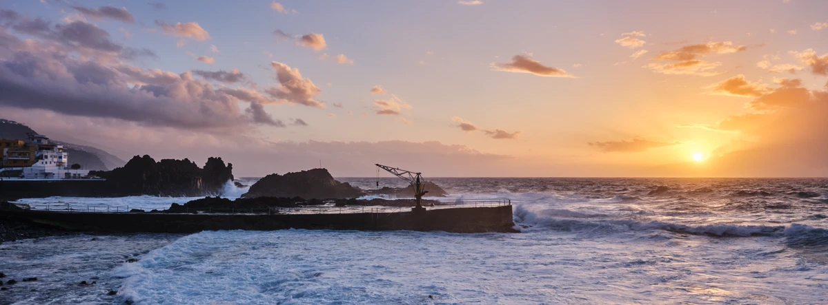 El Pris au coucher du soleil, rayons filtrant à travers les nuages, vagues à l'assaut des rochers.