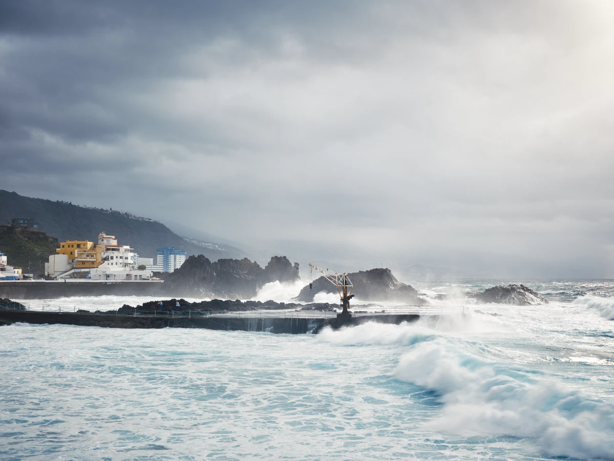 Vagues de tempête s'écrasant devant des façades ocre et jaune de El Pris, ciel bas et menaçant.